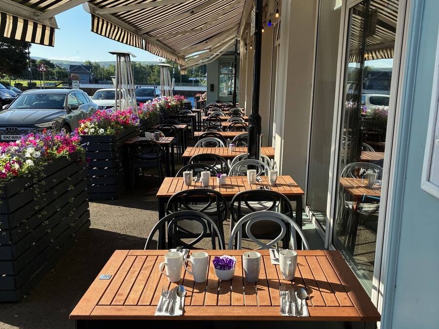 An outdoor cafe with wooden tables and chairs, a striped awning, and flower planters.