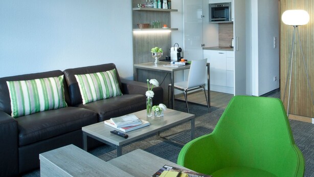 Un salon moderne avec un canapé, un fauteuil vert, une table basse et, au fond, un coin cuisine.