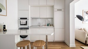 Una cocina moderna con muebles blancos, horno empotrado, lavadora y un comedor con taburetes.