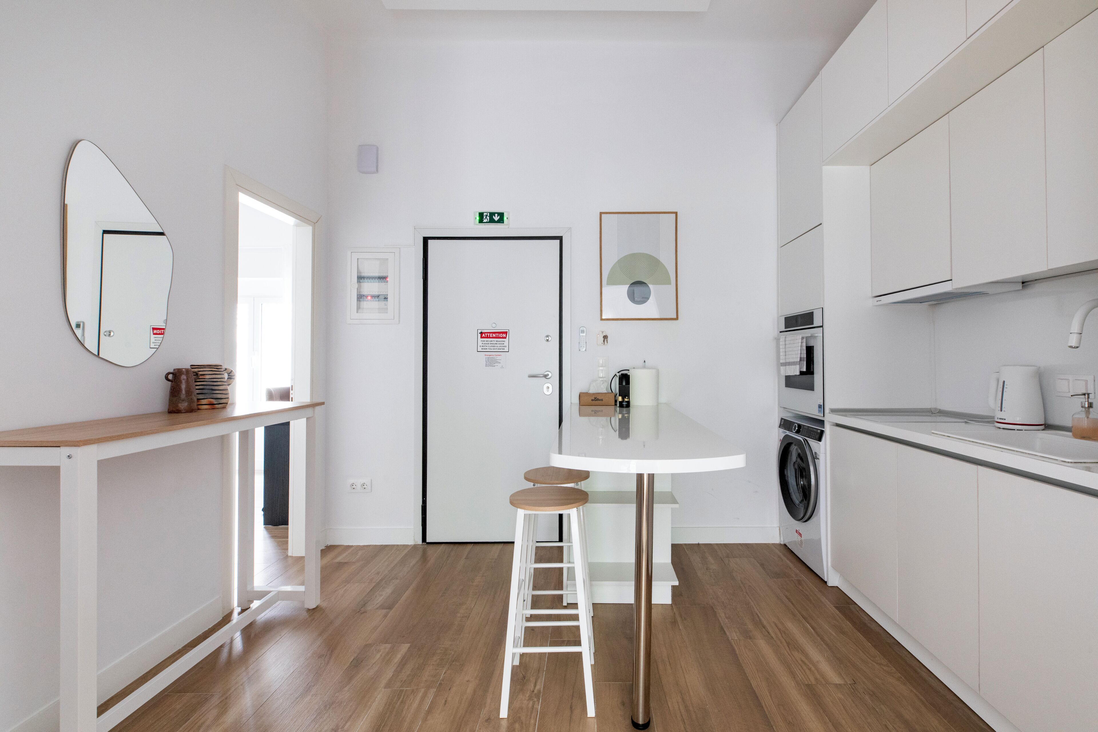 Une cuisine moderne équipée d’un lave-linge, d’une petite table avec des tabourets et d’une porte portant un panneau indiquant la sortie de secours.