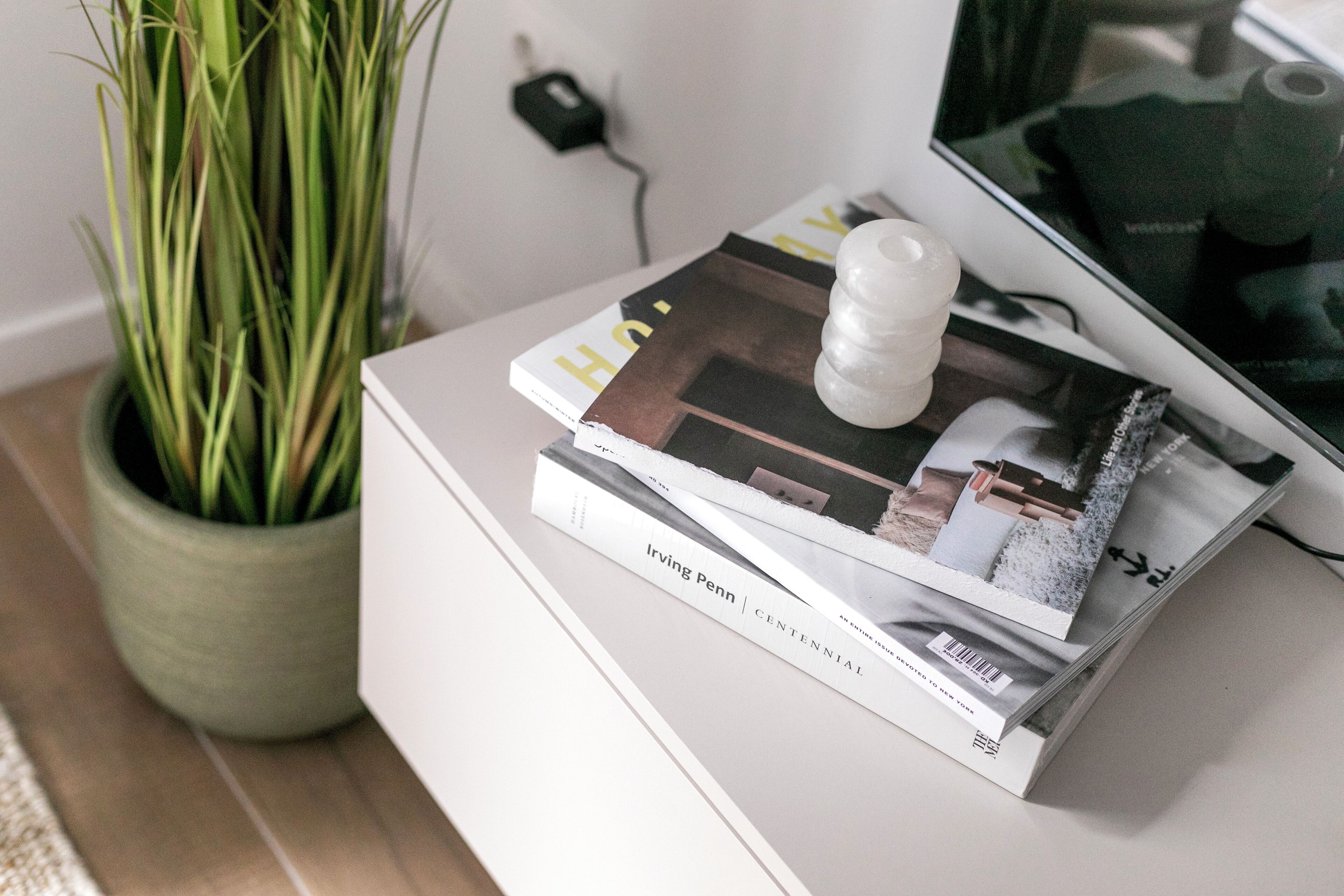 A stack of magazines on a white table with a potted plant beside it.