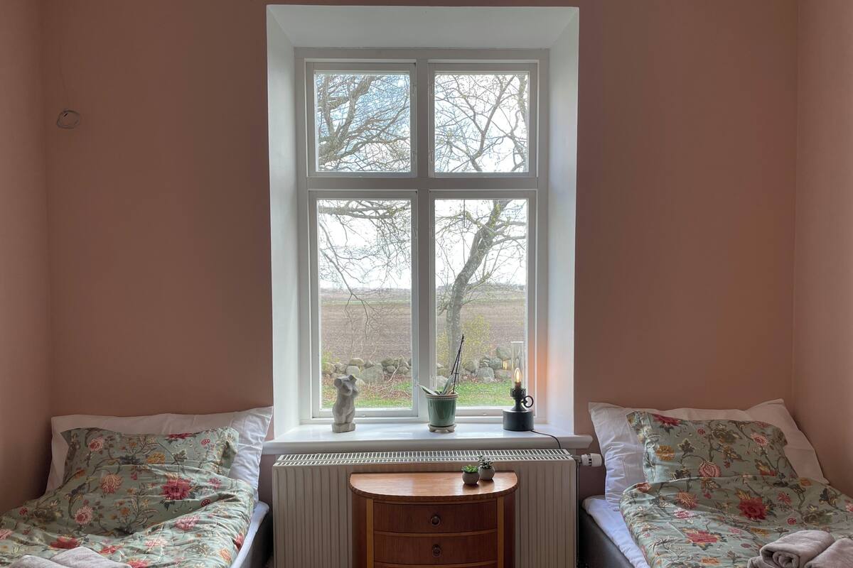 A room with a large window, a radiator, a wooden table, and two beds with floral bedding.