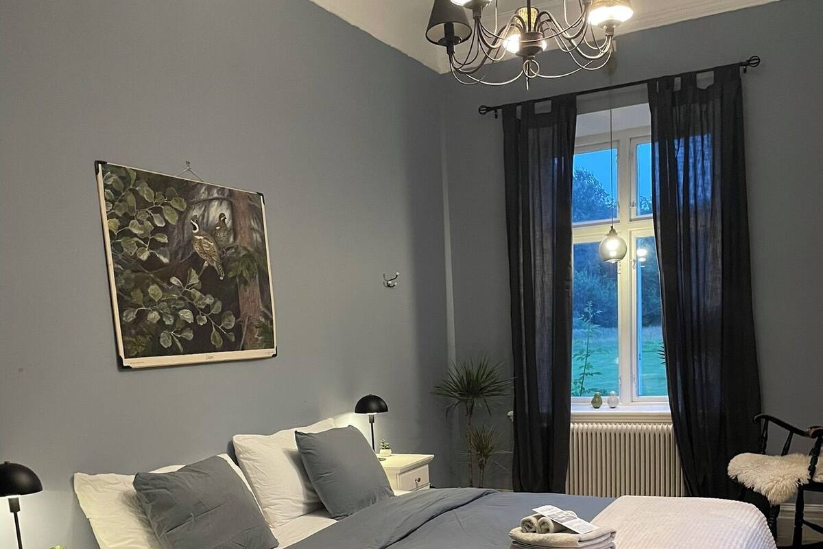 A bedroom with a bed, bedside tables, a nightstand, a window with curtains, a chandelier, and a wall-mounted painting.