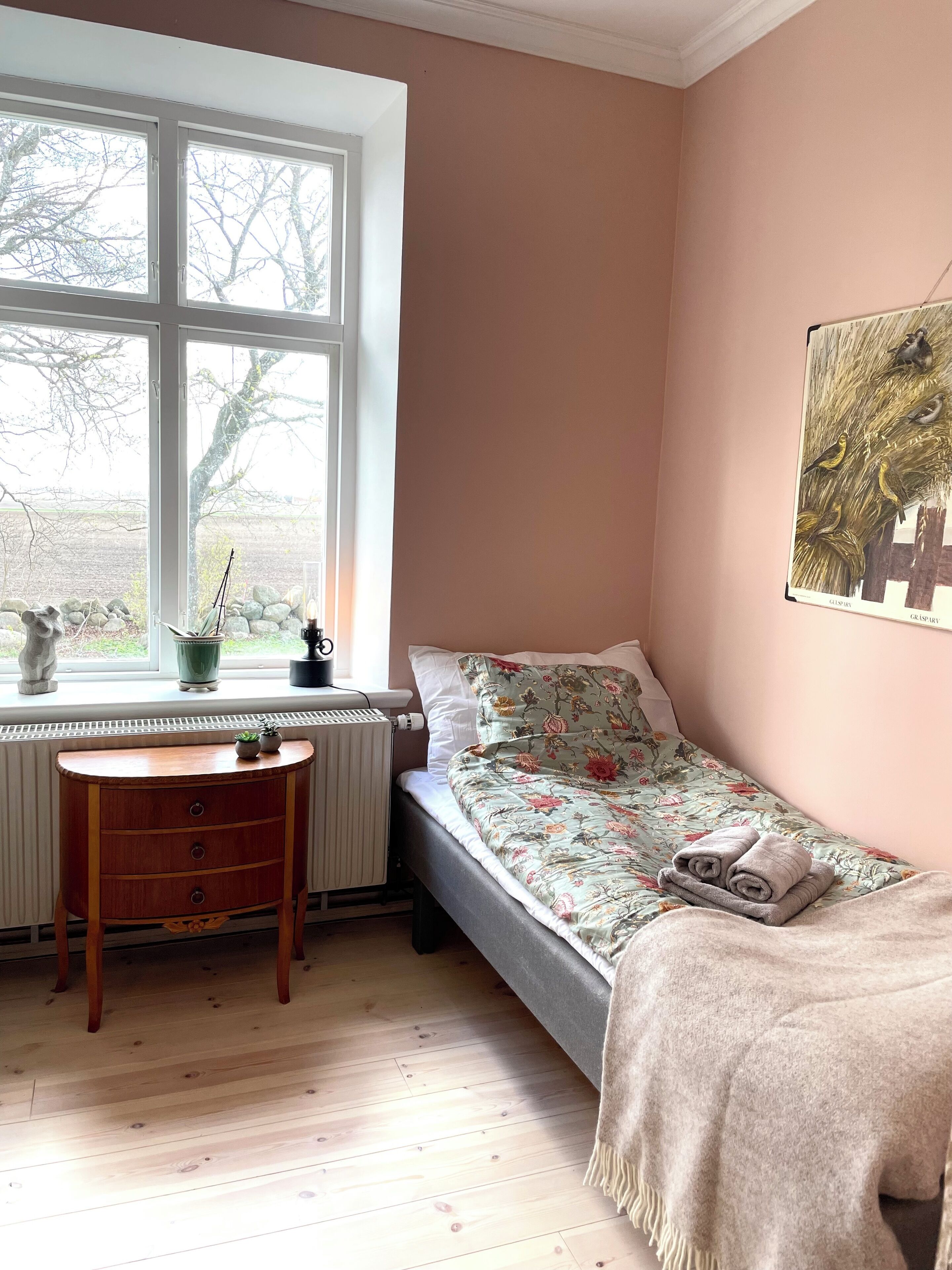 A small bedroom with a single bed, a radiator, a window with a view of trees, a small table with a plant, and a painting on the wall.