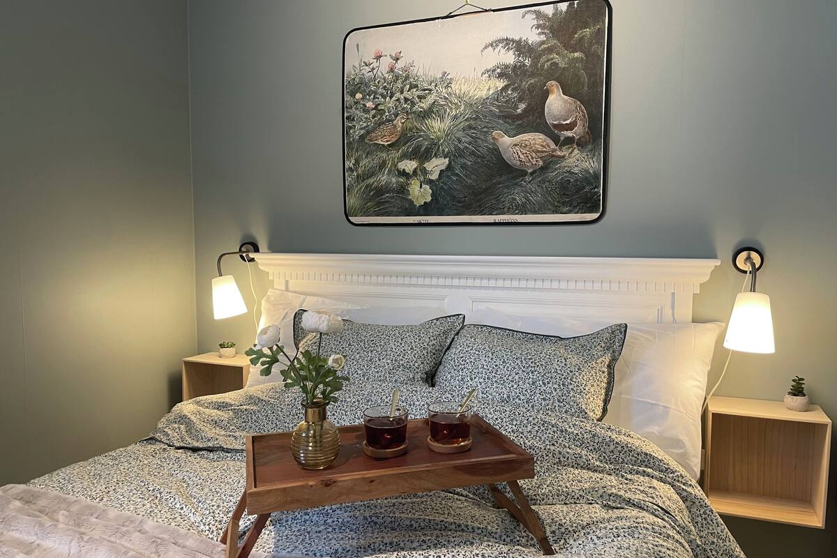 A bedroom with a bed, a bedside table, a tray with a vase and glasses, and a painting on the wall.