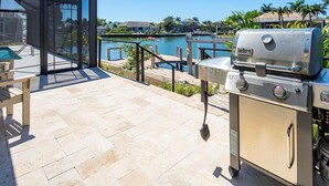 Een terras buiten met een barbecue, een glazen omheining en uitzicht op een waterpartij.