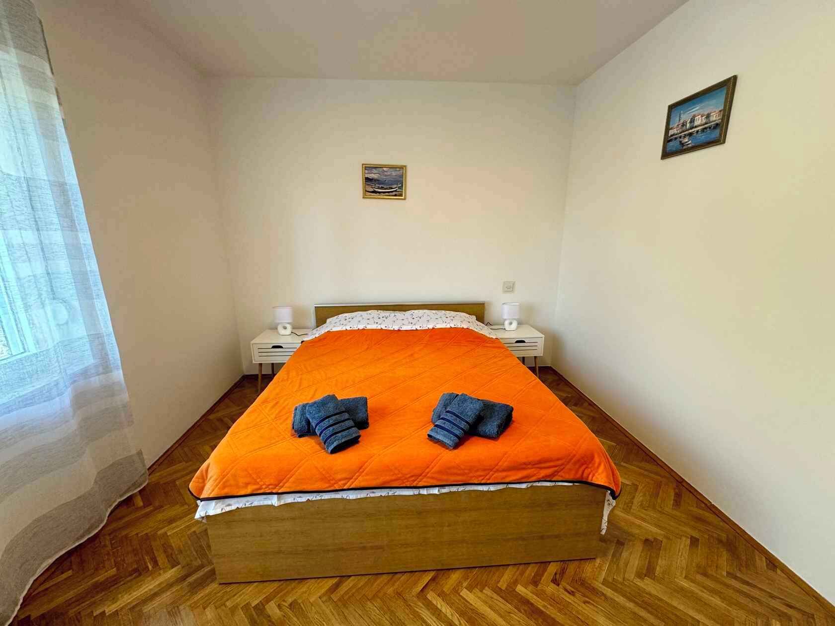 A bedroom with a wooden bed, an orange bedspread, two towels, a nightstand, a lamp, and a small painting on the wall.