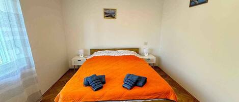 A bedroom with a wooden bed, an orange bedspread, two towels, a nightstand, a lamp, and a small painting on the wall.