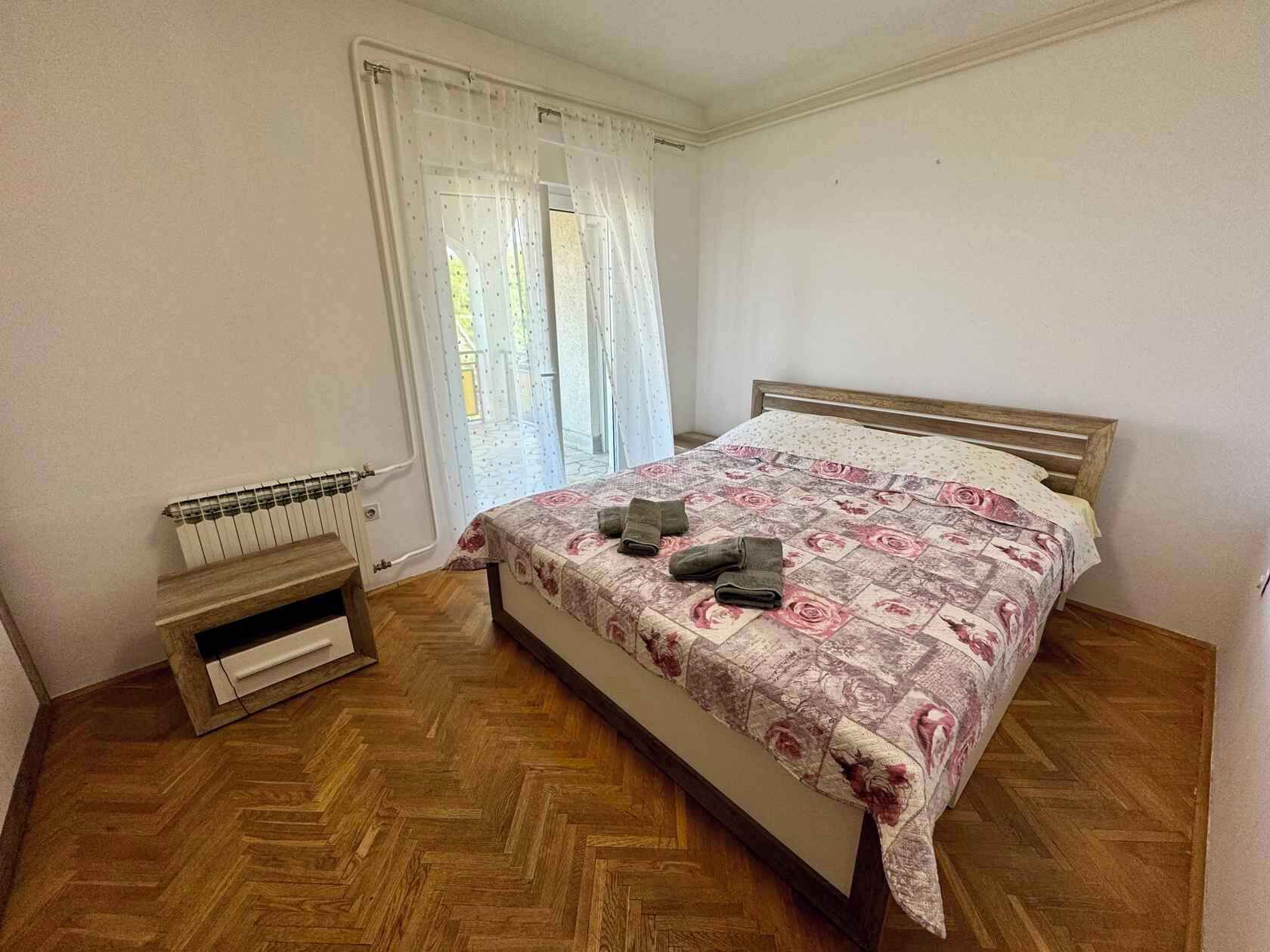 A bedroom with a wooden herringbone floor, a bed with a floral bedspread, a radiator, and a window with sheer curtains.