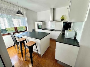 Kitchen features a sleek island with a black countertop, white cabinetry, oven, microwave, and range hood, with a large window offering a view of the outdoors.