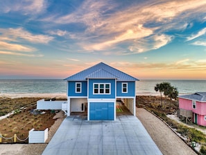 Uma casa de praia de dois andares com fachada azul e porta de garagem branca, situada numa praia de areia e vista para o oceano.