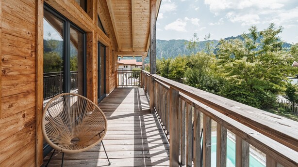 A wooden balcony with a wicker chair, large windows, and a view of a mountainous landscape. - 6 persons Chalet Bergherzerl in the Chiemgau. Summer pool, whirlpool & sauna (Inzell)