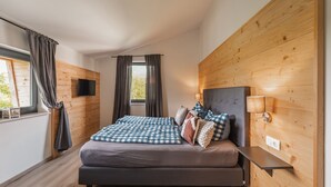 A modern bedroom with a large bed, wooden panel walls, a flat-screen TV, and a bedside table with a lamp. - 6 persons Chalet Bergherzerl in the Chiemgau. Summer pool, whirlpool & sauna (Inzell)