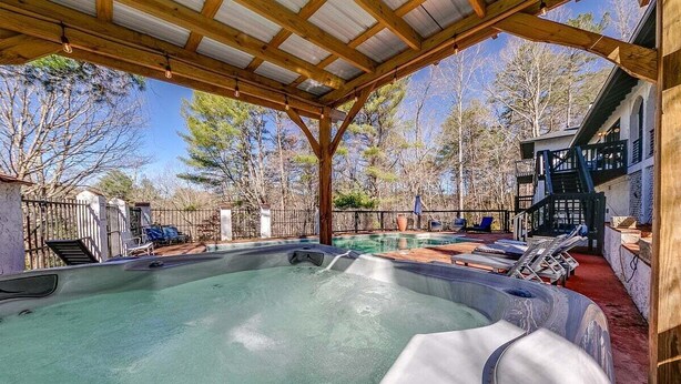 A hot tub under a wooden pergola with a pool in the background, surrounded by trees and a fenced area.