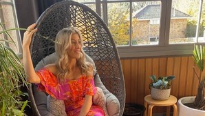 A person sitting in a wicker chair on a tiled patio with potted plants and a view of houses through large windows.