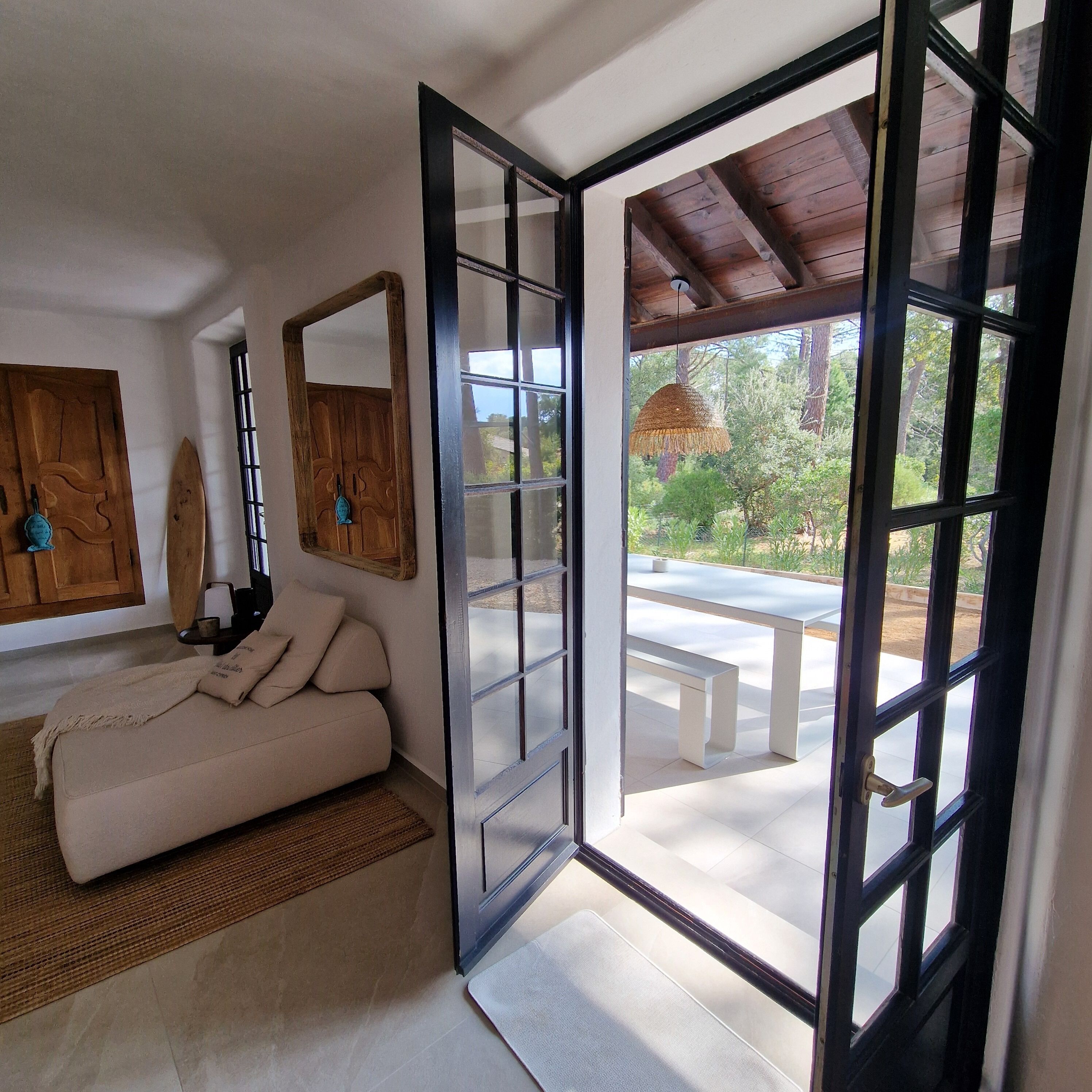 A room with a sofa, a wooden door, a mirror, and a view of an outdoor patio with a table and bench.
