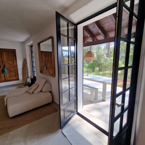A room with a sofa, a wooden door, a mirror, and a view of an outdoor patio with a table and bench.