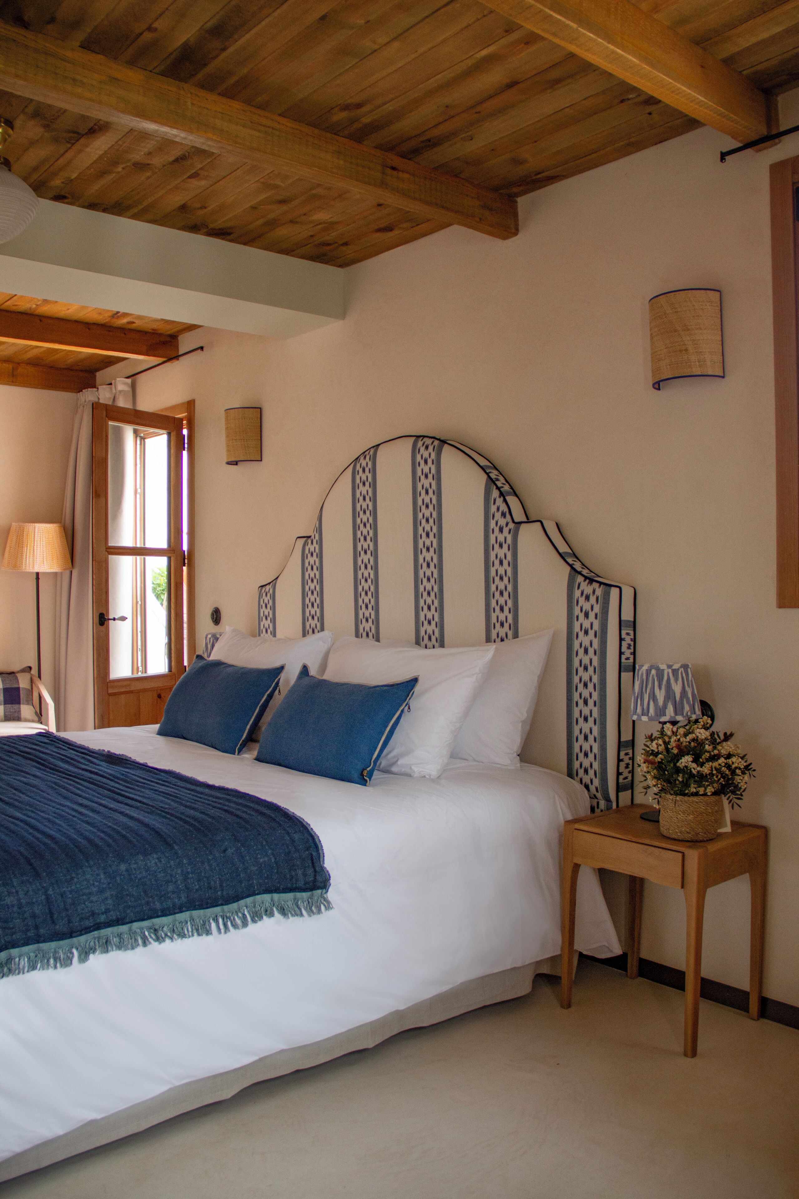 Un dormitorio con techo de madera, una cama con ropa de cama blanca y azul, una mesita de noche con una lámpara y una ventana con cortinas.