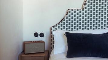 Una cama bien tendida con un cabecero estampado, una mesita de noche de madera pequeña con una radio de estilo vintage, y un cojín de color oscuro.