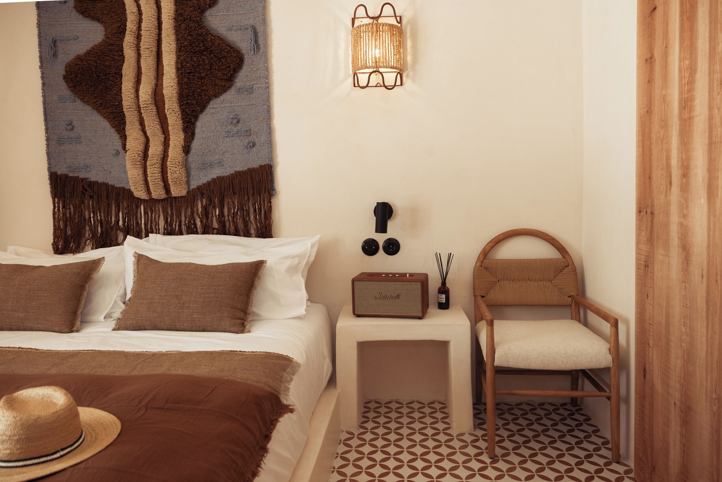 A bedroom with a bed, a nightstand with a lamp, a chair, and a wall tapestry.