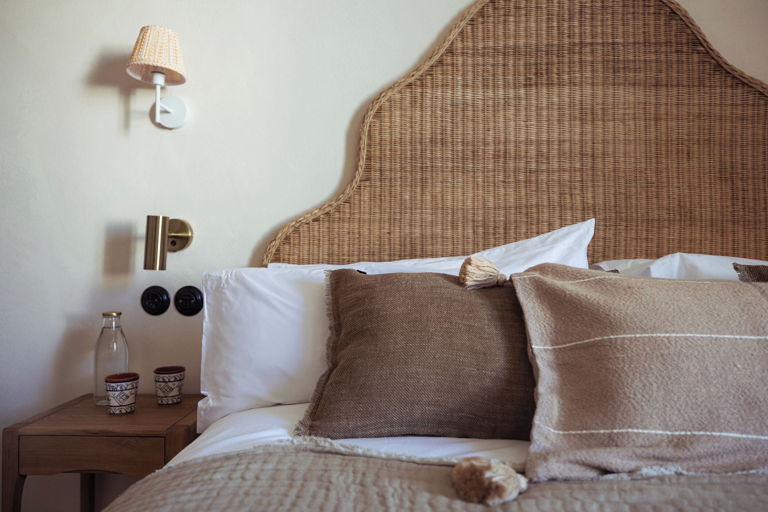 A neatly made bed with a woven headboard, a bedside lamp, and two decorative vases on a wooden nightstand.