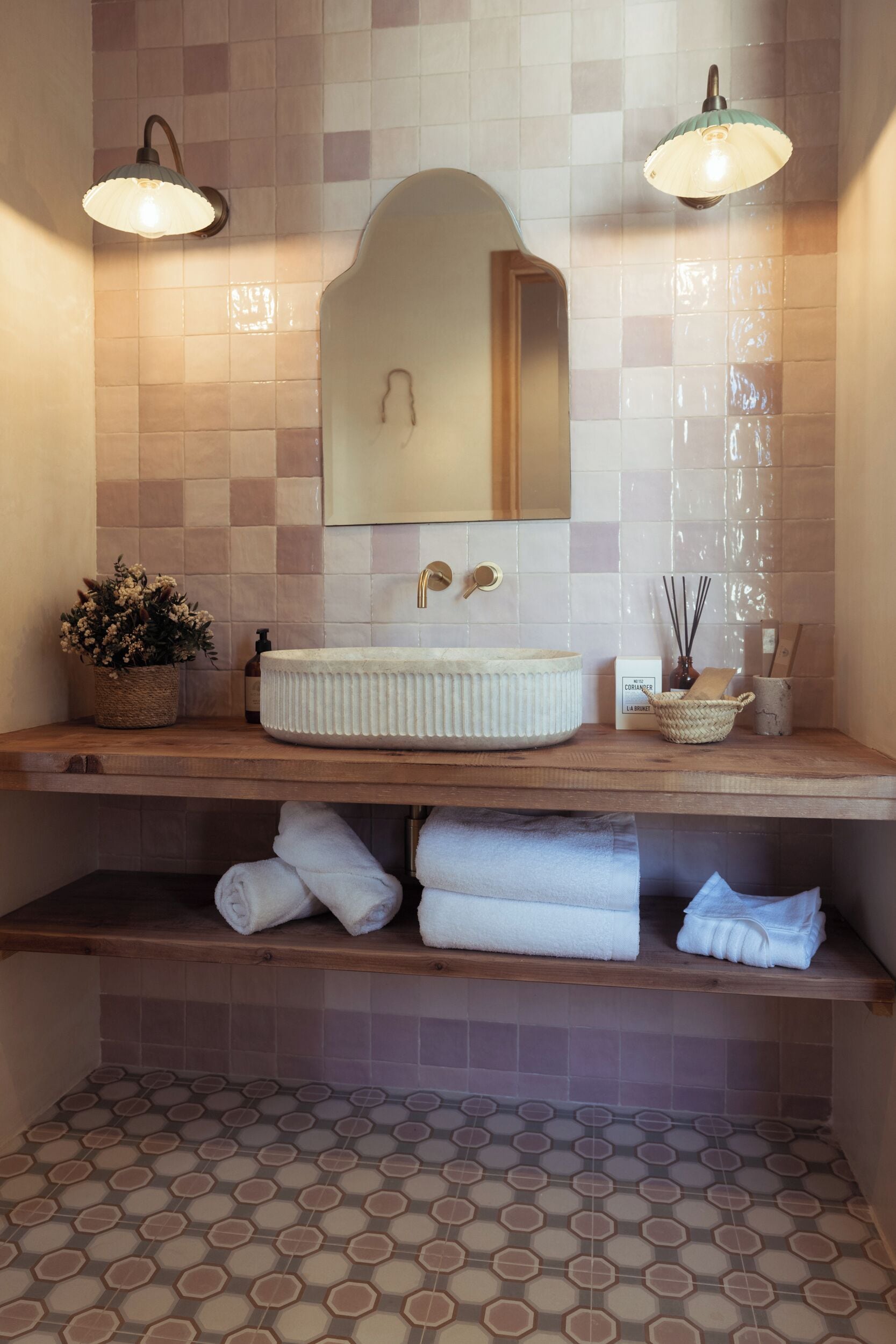 A bathroom with a white sink, two wall-mounted lights, a mirror, a potted plant, and neatly folded towels on a shelf.
