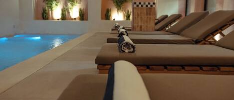 Un área junto a la piscina con sillones de descanso, una piscina pequeña y ventanas arqueadas con plantas.