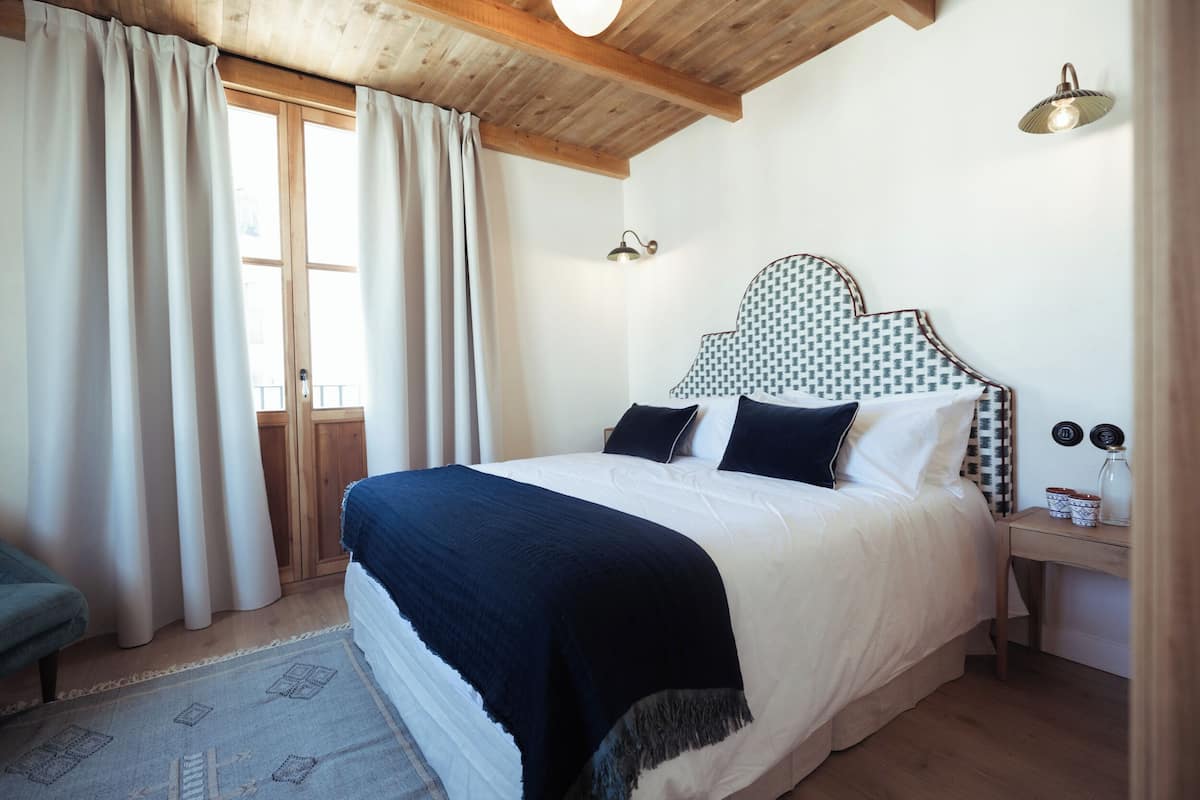 A bedroom with a large bed, a wooden ceiling, a nightstand with a lamp, and a window with curtains.