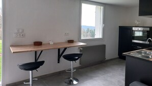 A modern kitchen with a wooden bar counter, two black bar stools, a built-in oven, and a window providing a view of the outdoors.