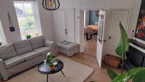 Un salon avec un canapé, une table basse et des plantes. On aperçoit un coin repas avec une table et des chaises à travers une porte ouverte.