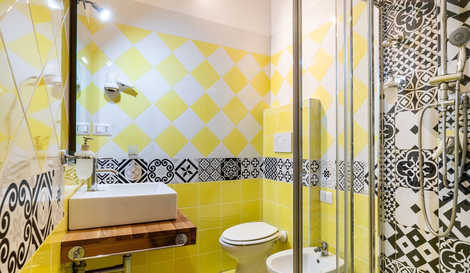 A bathroom with a yellow and white tile pattern, a wooden vanity, a sink, a toilet, and a shower area with patterned tiles.