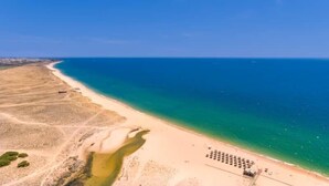 Vista aerea di una spiaggia con cielo azzurro limpido, un litorale sabbioso e alcuni veicoli parcheggiati.