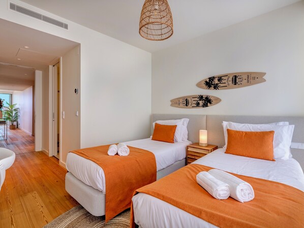 A hotel room with two beds, each with an orange blanket and white pillows, a bedside table with a lamp, and a wall with surfboard decorations.