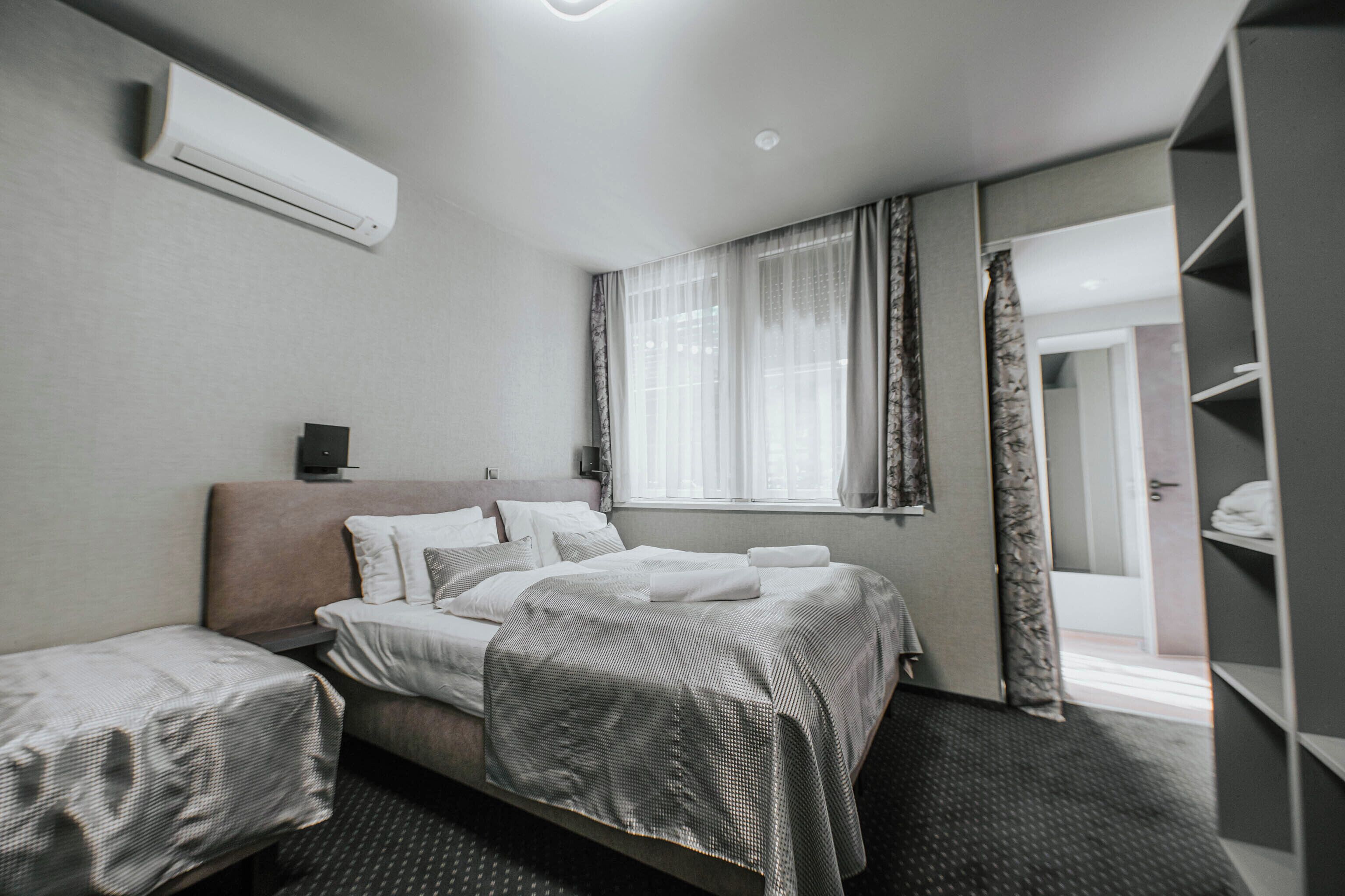 A hotel room with a bed, a bedside table, an air conditioner, a window with curtains, and a wardrobe.
