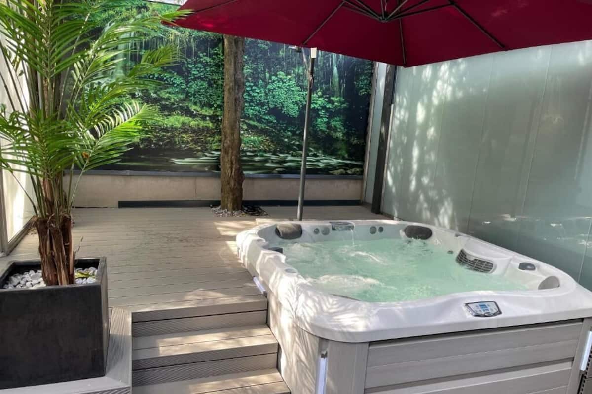 A hot tub with a view of a forested area, a red umbrella, and a potted plant.