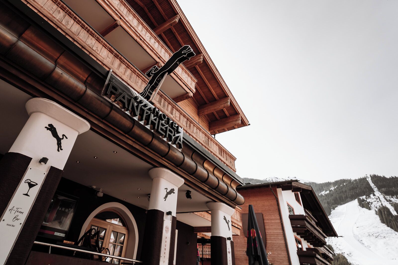 A hotel entrance with a wooden facade, a sign reading "Hotel Antlher," and a ski slope in the background.