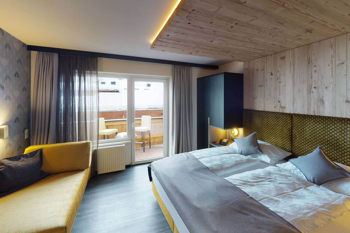 A hotel room with a large bed, a sofa, a balcony with a view, and a wooden ceiling.