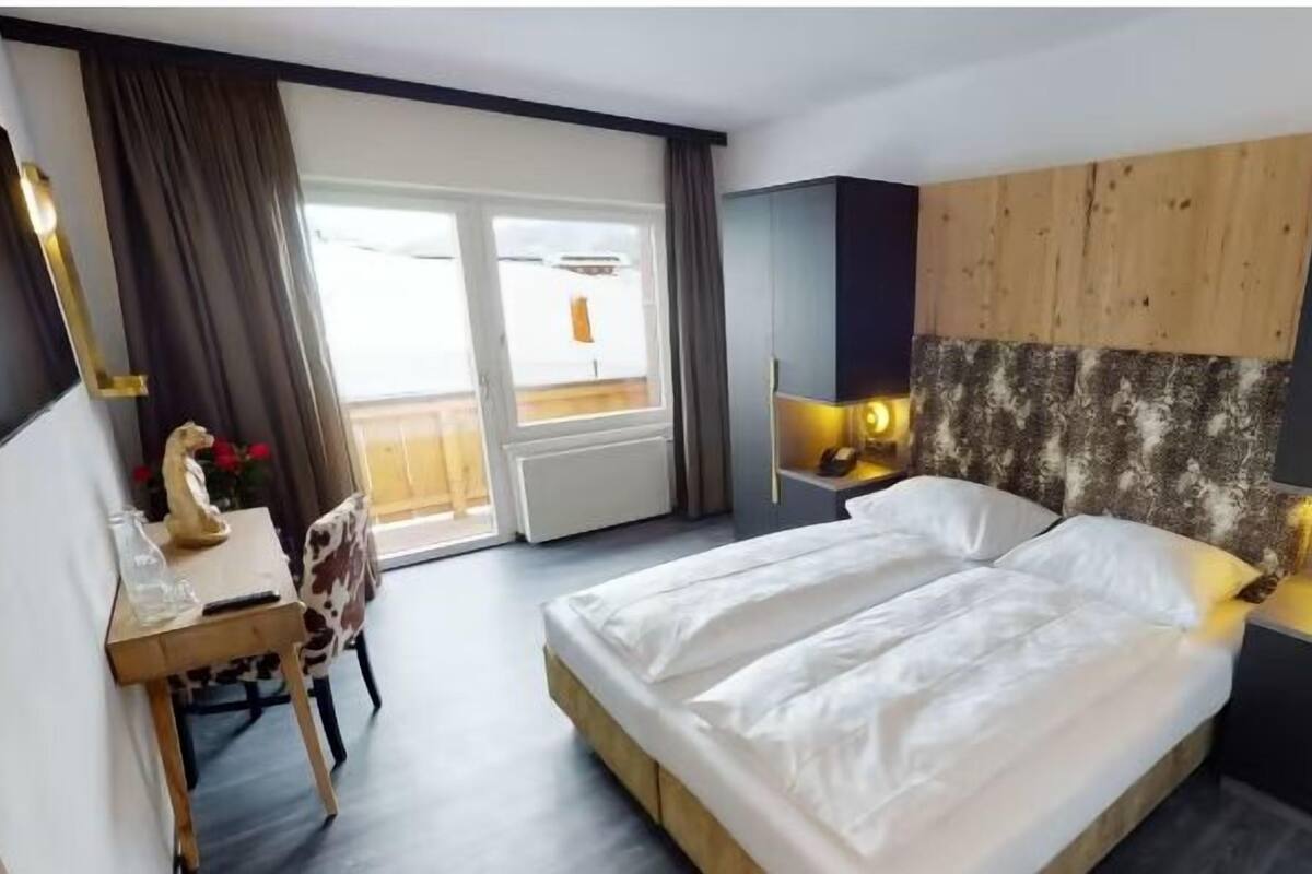 A hotel room with a bed, a desk, a chair, a wardrobe, and a view of the outside.