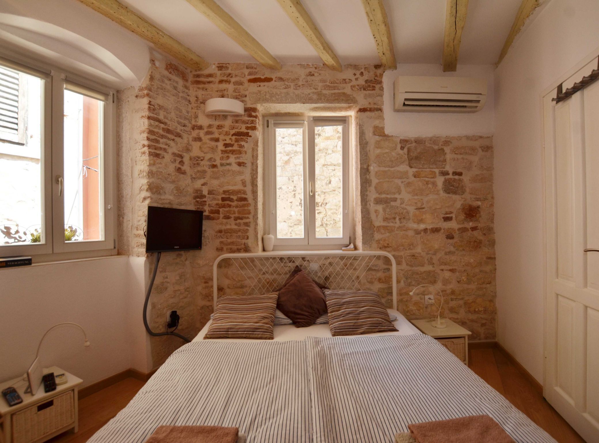 A bedroom with a stone wall, a bed with striped bedding, a TV mounted on the wall, and a window with a view of the outside.