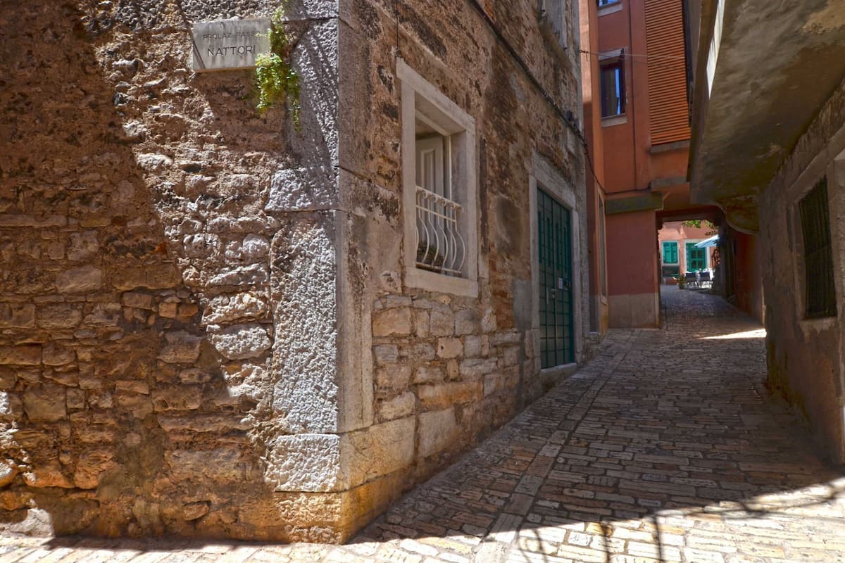 A narrow cobblestone street with stone buildings, a sign reading "NATTIORI," and a window with metal bars.
