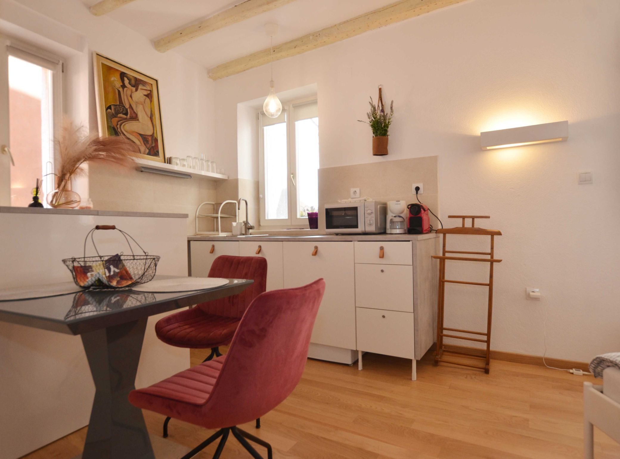 A modern kitchen with white cabinets, a microwave, a coffee maker, and a hanging plant. There is a dining table with two red chairs and a basket of fruit.