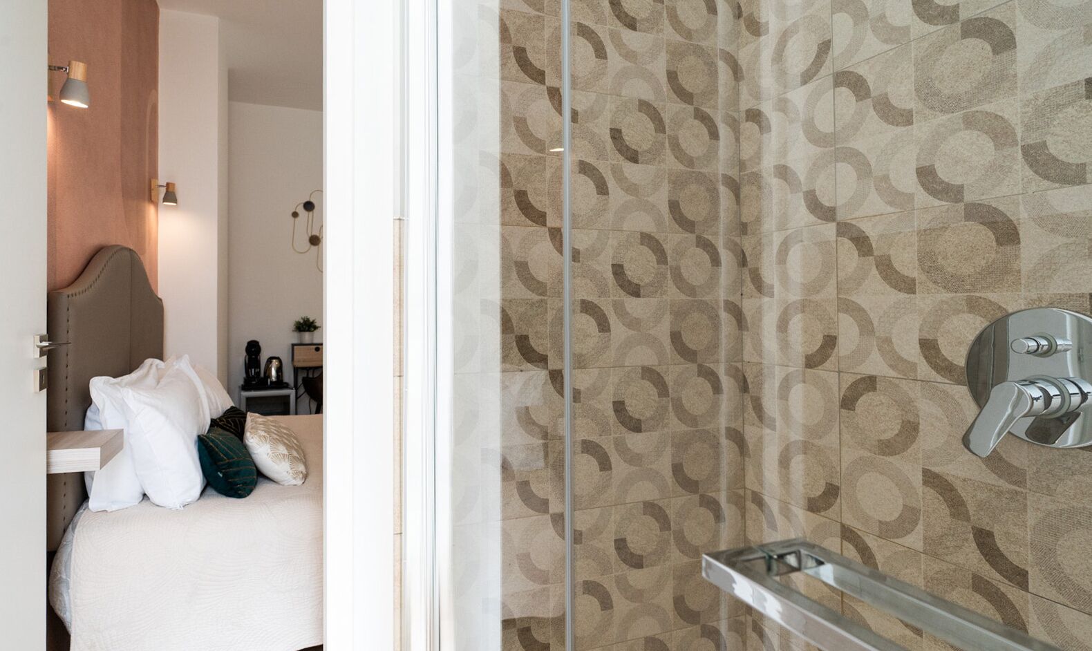 A bathroom with patterned tiles, a shower with a chrome faucet, and a bed with pillows.
