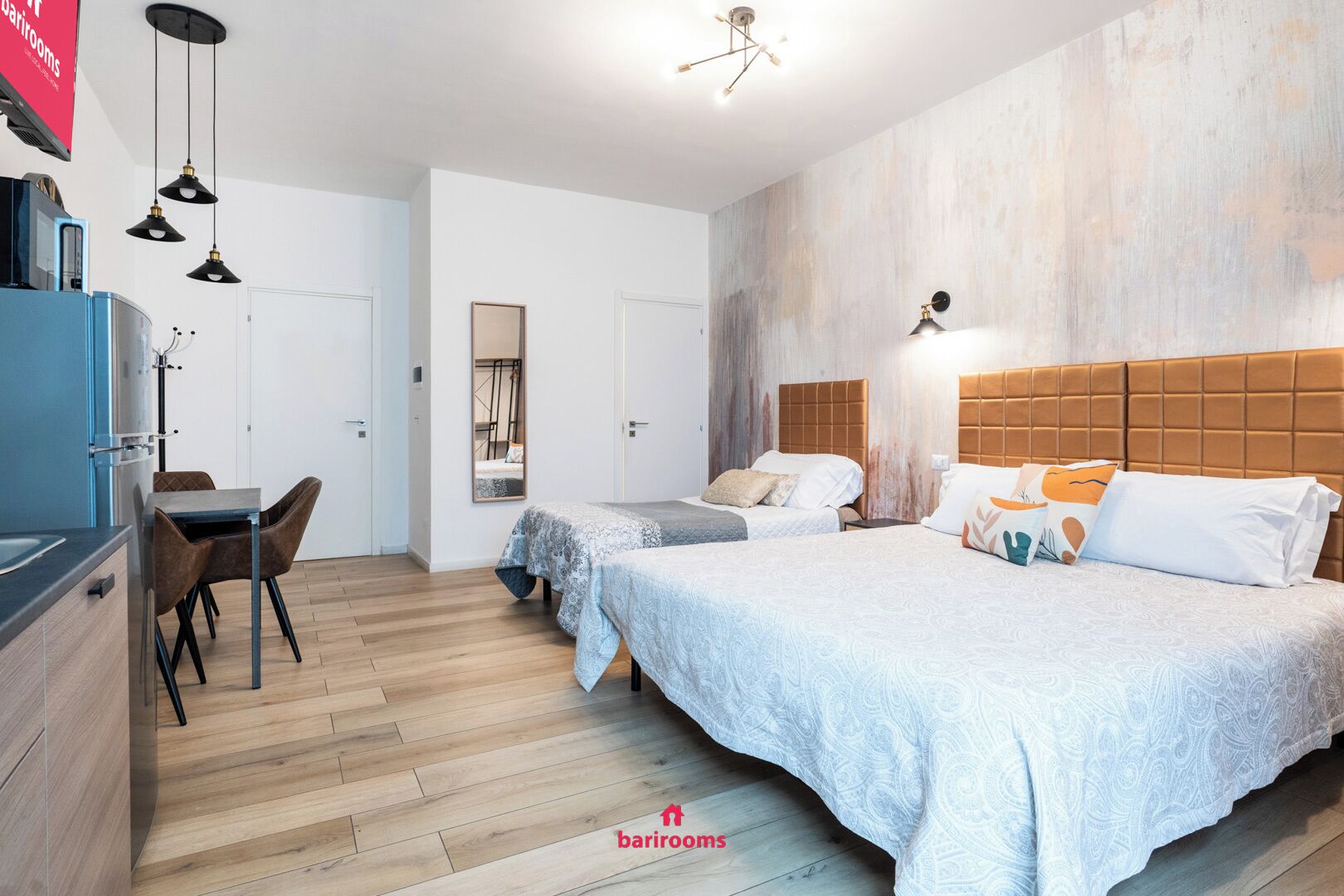 A modern hotel room with a wooden floor, two beds with white linens, a dining area with a table and chairs, and a kitchenette.