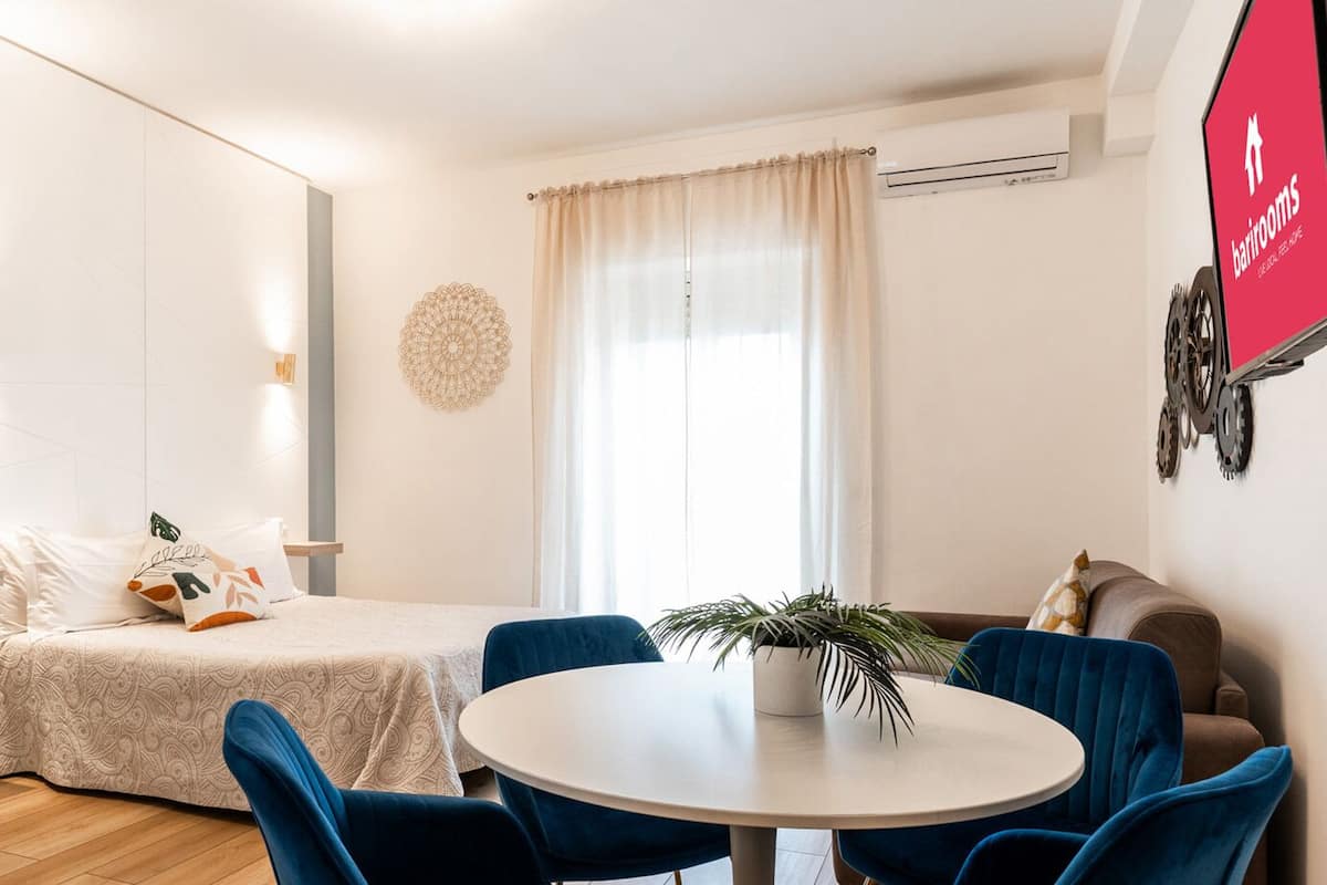 Ein modernes Hotelzimmer mit einem Bett, einem runden Esstisch, blauen Stühlen, einer Topfpflanze und einer Wand-Klimaanlage.