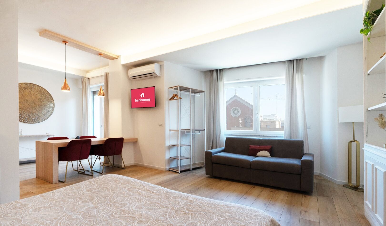 A modern hotel room with a bed, a sofa, a dining area with chairs, a TV, and a window with curtains.