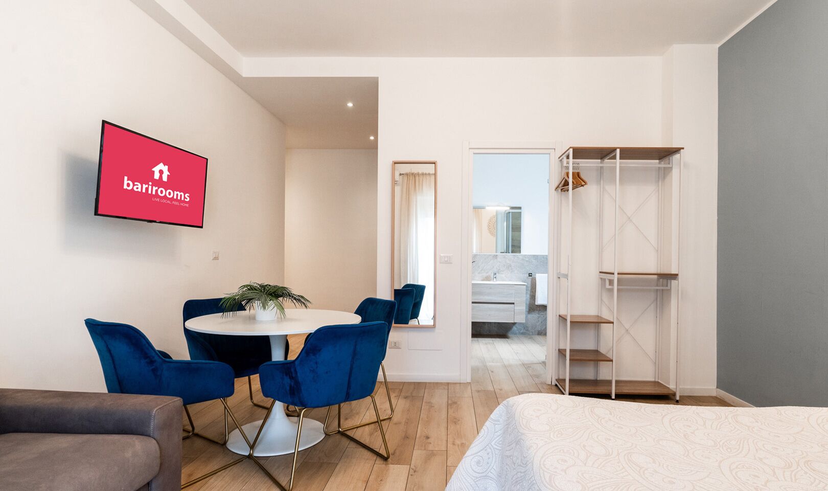 A modern hotel room with a round dining table, blue chairs, a grey sofa, a bed with a patterned duvet, and a TV displaying the barrooms logo.