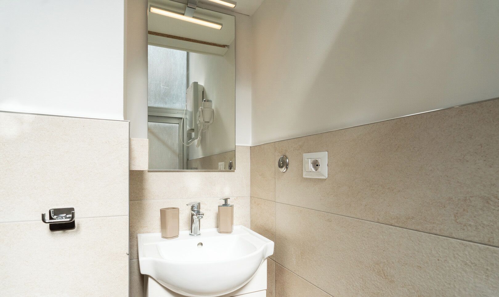 A bathroom with a white sink, two soap dispensers, a mirror, and a shower visible in the reflection.