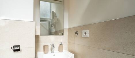 Un baño con un lavamanos blanco, dos dispensadores de jabón, un espejo y una ducha visible en el reflejo.