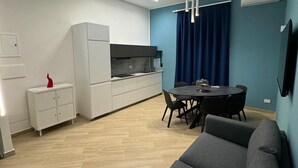 Un espacio habitable moderno con un sofá gris, una mesa de comedor con sillas, una zona de cocina con armarios blancos y una pared azul de acento.