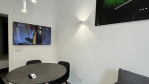A modern room with a round table, black chairs, a grey sofa, a flat-screen TV mounted on the wall, and a ceiling light fixture.